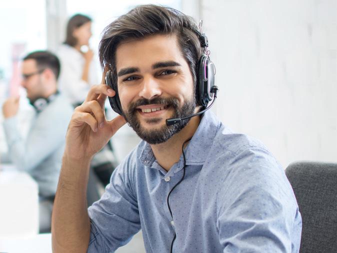 Junger Mann mit Headset am Schreibtisch l&auml;chelt in die Kamera, w&auml;hrend er an einer Tastatur arbeitet.