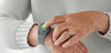 Nahaufnahme einer Hand, die einen gr&uuml;nen Knopf auf einem grauen Armband dr&uuml;ckt, w&auml;hrend eine Person einen grauen Pullover tr&auml;gt.