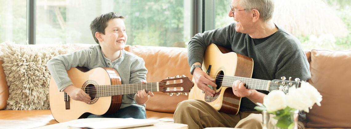 Junge und &auml;lterer Mann spielen gemeinsam Gitarre auf einer Couch, umgeben von Pflanzen und einem Blumenstrau&szlig;.