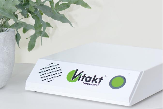 Hausnotrufger&auml;t von Vitakt f&uuml;r den Notruf zuhause, neben einer grauen Pflanze im Topf.