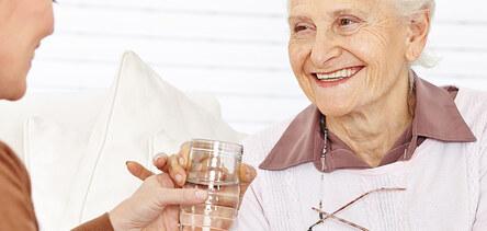 L&auml;chelnde &auml;ltere Frau erh&auml;lt ein Glas Wasser von einer j&uuml;ngeren Person in einem hellen Raum.