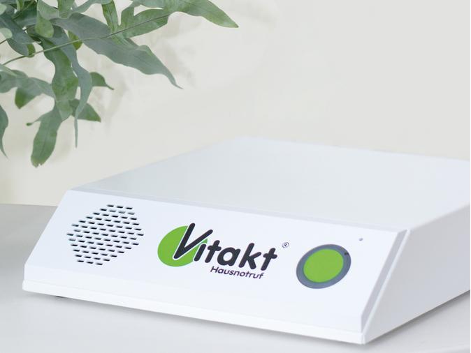 Hausnotrufger&auml;t von Vitakt f&uuml;r den Notruf zuhause, neben einer grauen Pflanze im Topf.