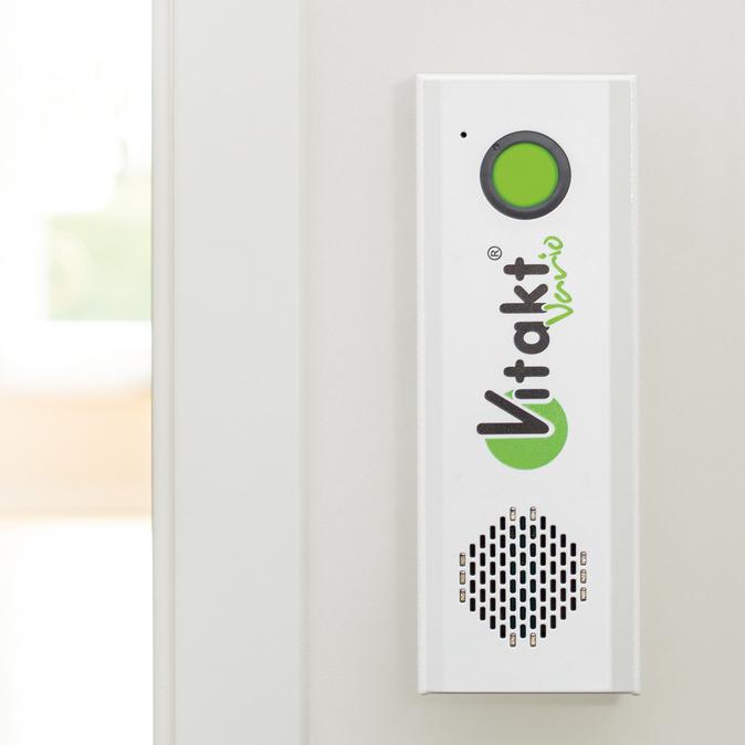 Wei&szlig;es Ger&auml;t mit gr&uuml;ner Taste und Lautsprecher, an einer Wand in einem hellen Raum montiert.