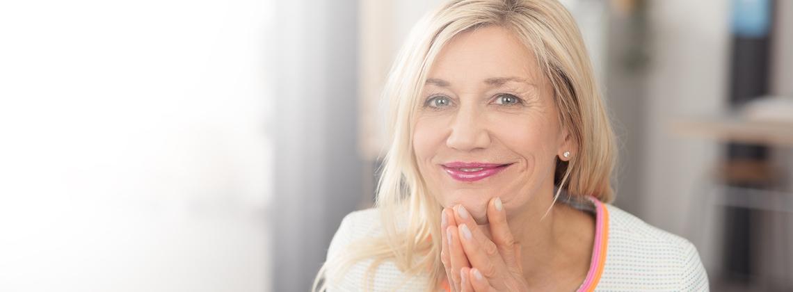 Frau mit blonden Haaren l&auml;chelt in die Kamera, H&auml;nde unter dem Kinn gest&uuml;tzt, in einem hellen Innenraum.