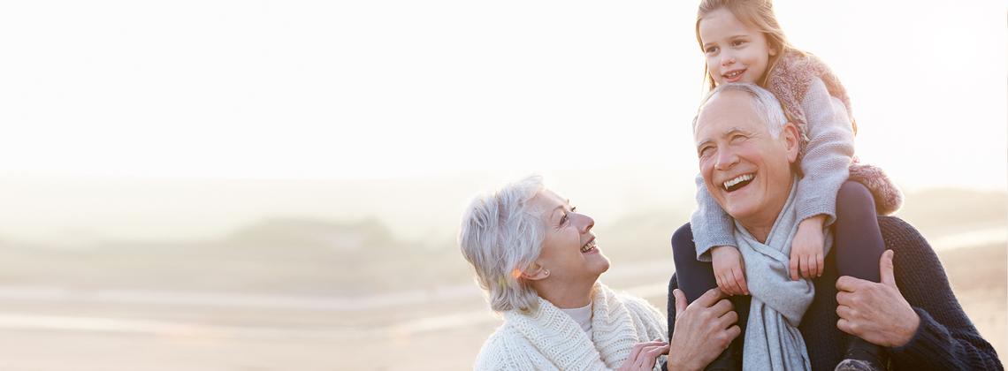 Ein &auml;lteres Paar lacht, w&auml;hrend ein Kind auf dem R&uuml;cken des Mannes sitzt, vor einer hellen, unscharfen Landschaft.